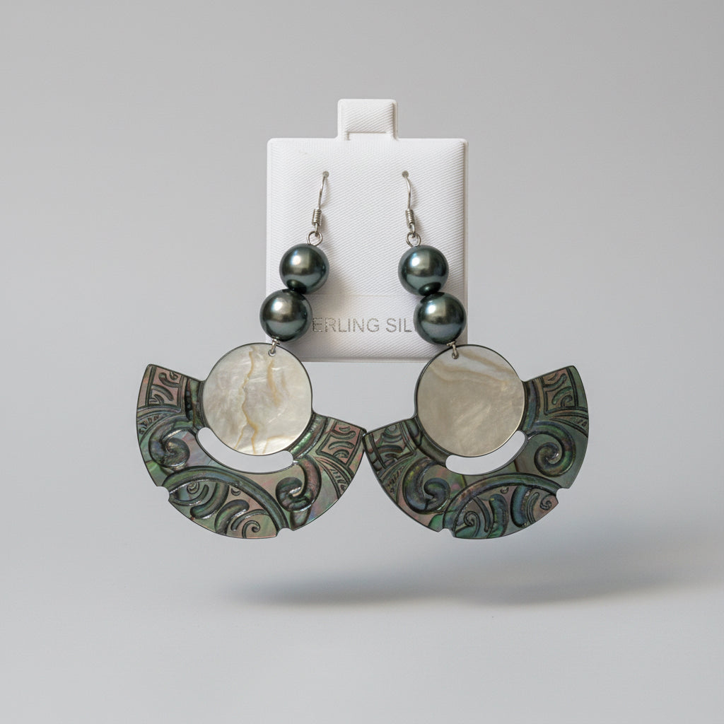 A pair of dangling earrings displayed on a white card, resting on a woven straw mat with yellow-and-white flower petals at the top. The earrings have dark gray pearl-like round beads and silver-toned metal hooks, with large iridescent shell disc and scroll-shaped pieces in green, blue, and silver tones.